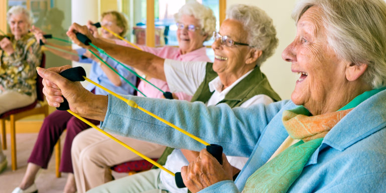 Large group of happy enthusiastic elderly ladies exercising in a gym sitting in chairs doing stretching exercises with rubber bands.